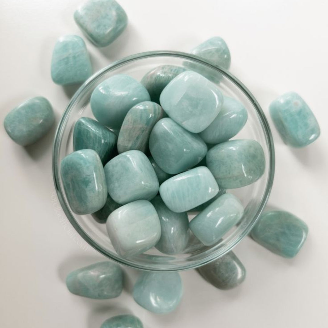 Smooth turquoise-green amazonite stones piled in a clear glass bowl, with additional stones scattered around the outside.